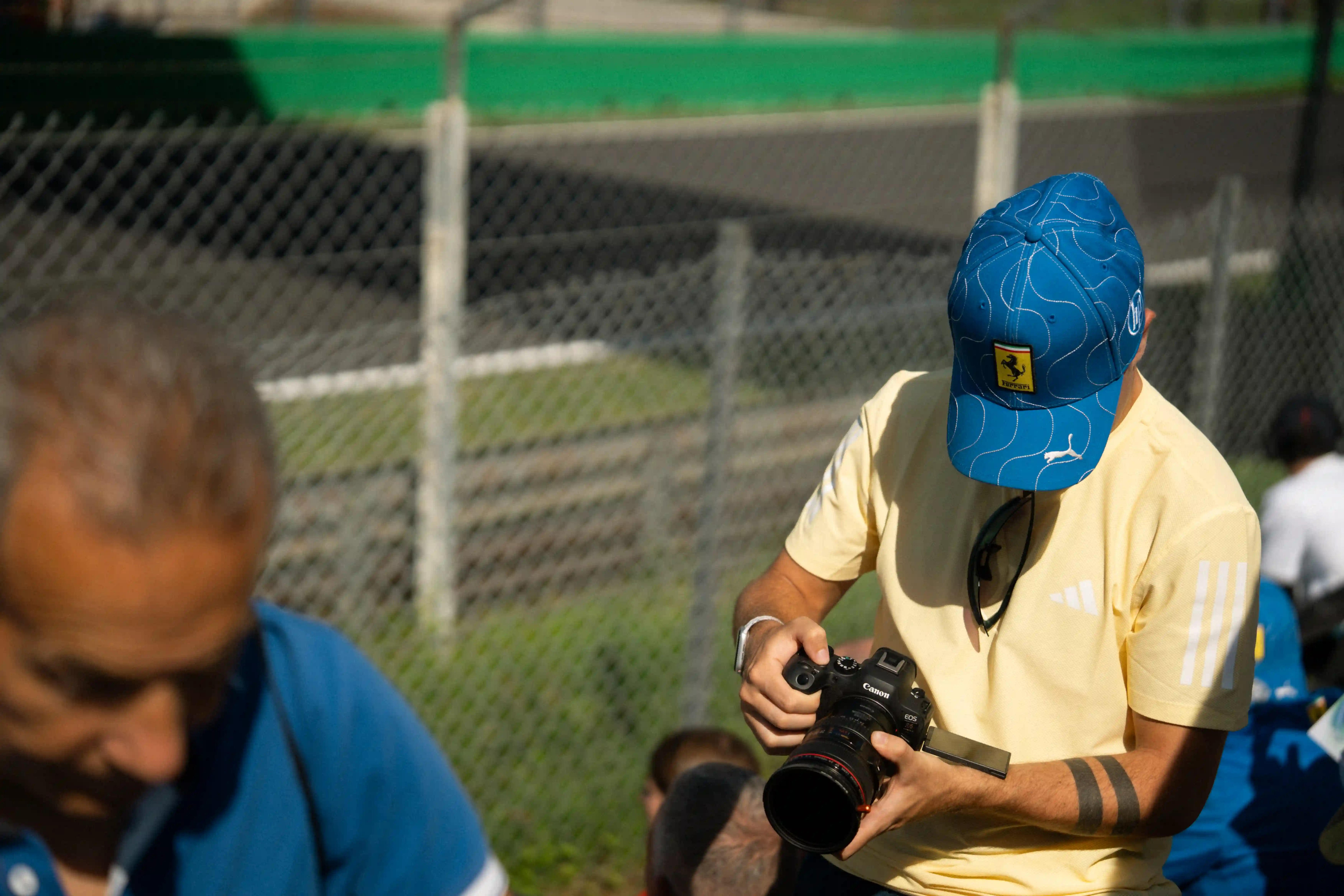 Man in yellow shirt and blue Ferrari hat holds camera.